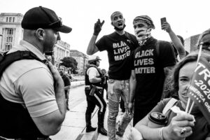 Man in BLM shirt shouts at cop while Trump lady peers out from flag