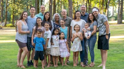 Portrait of a senior couple posing for a photograph with their adult children and grandchildren. The multi-ethnic and multi-generation group is gathered at a family reunion. The family members affectionately have their arms around one another and are smiling. They are standing in a grassy meadow with trees in the background. Stock photo