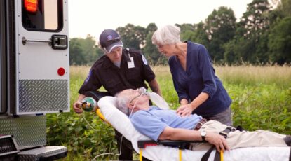 Elderly patient lays on a gurney while elderly companion comforts him and EMT loads him into waiting ambulance.