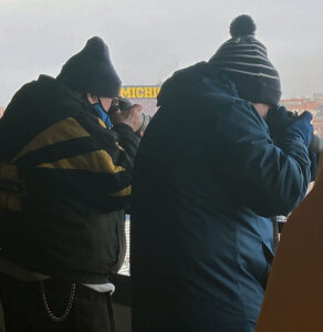 Dick Gaskill and Mark Gaskill, dressed head to toe in winter/U-M gear, stand side by side, shot from behind, as they take pictures on the field at Michigan Stadium.