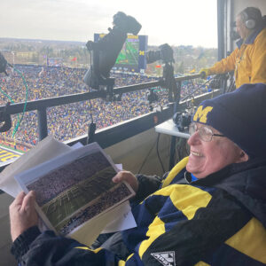 Gaskill, dressed head to toe in U-M gear sits high atop Michigan Stadium holding his favorite photo. You can see the crowd in the distance.