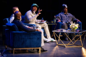 Seated African American man wearing ballcap addresses an unseen audience with hand gesture.