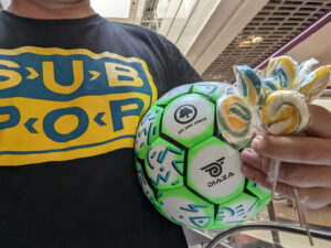 Kid with Sub Pop t-shirt holds soccer ball that reads AFC Ann Arbor and a handful of blue & yellow lollipops.