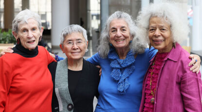 Four beautiful elderly women of various races dressed in bright colors and smiling with their arms around each other.