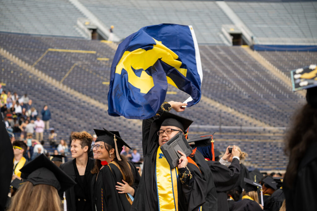 Grad in cap and gown waves Michigan flag in Michigan Stadium.