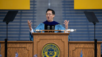 President Ono, an Asian male in glasses, stands at podium on graduation day. 2024