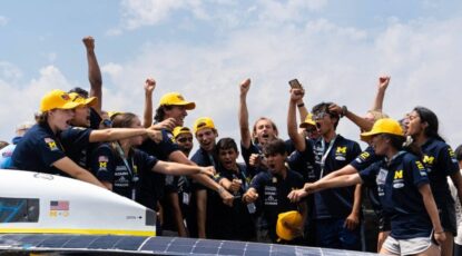 Members of U-M's 2024 champion Solar Car team surround the vehicle, dressed in blue shirts and maize caps with fists in the air.