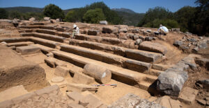 Archaeological field site features rocks, dirt, and crouching architects Kübra Sağlam and Gizem Seymen working on architectural drawings of Bouleuterion, view looking northwest.