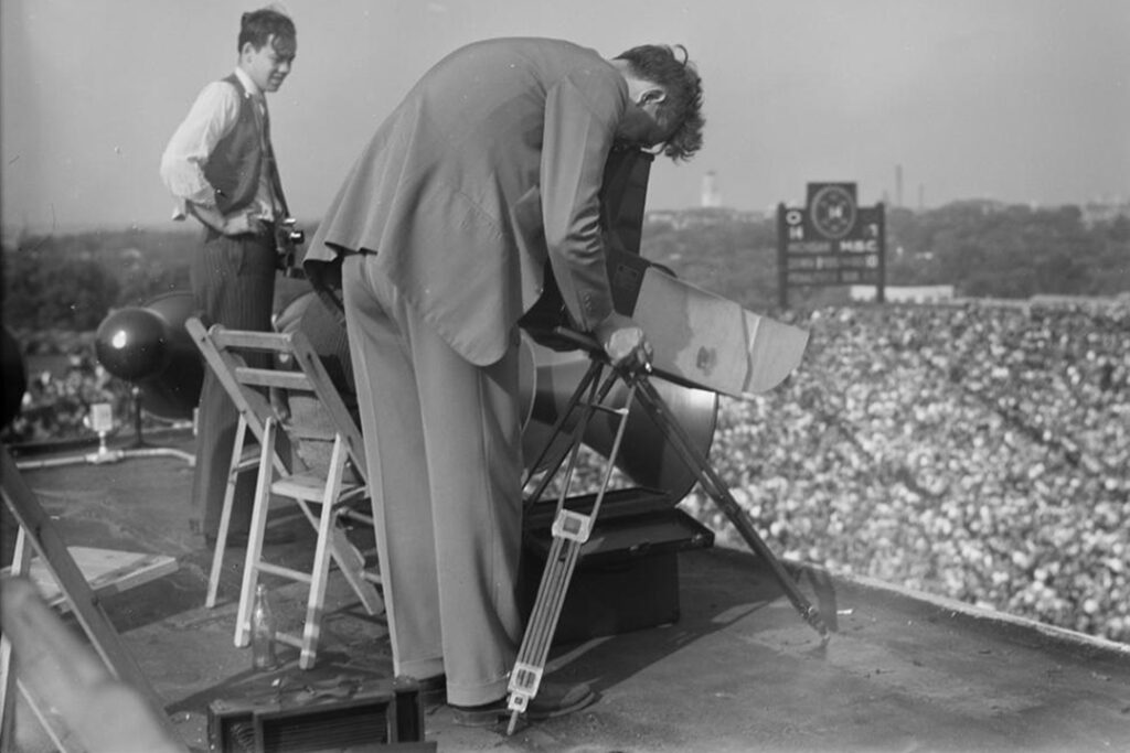 Man bends over camera circa 1940. Black & white image shows overview of Michigan Stadium.