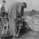 Man bends over camera circa 1940. Black & white image shows overview of Michigan Stadium.