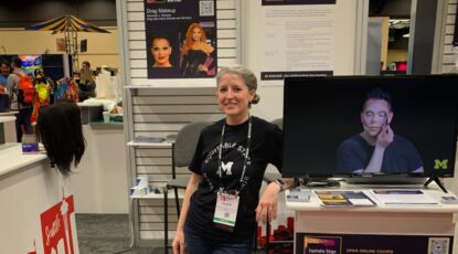 Woman stands next to video monitor that shows person applying stage makeup.