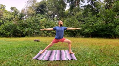 Man performing yoga -- warrior pose on striped rug on a grassy knoll.