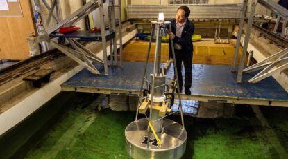 Four men stand on a platform stretching across a large indoor tank of water. A light shines on a metal buoy floating in the tank near the group. Lei Zuo (second right), a professor of naval architecture and marine engineering, Jeff Scruggs (second left), a professor of civil and environmental engineering, Xiaofan Li, an assistant research scientist of naval architecture and marine engineering (first right), and Jui-Chen Chen (first left), a masters student of naval architecture and marine engineering, demonstrate how a buoy can generate electricity at the University of Michigan’s Marine Hydrodynamics Laboratory. Image credit: Marcin Szczepanski, Michigan Engineering.