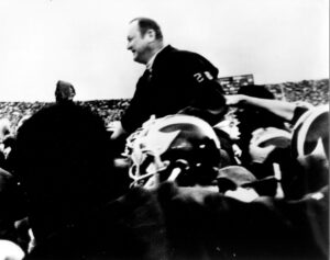 Black and white image of Michigan football coach Bo Schembechler on the shoulders of Wolverines who carry him off the field after November 1969 victory against Ohio State.