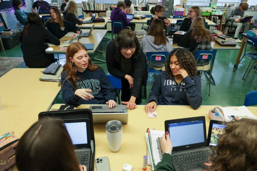 Ylisse Yépez, a teacher at Blissfield High School, stands between and talks to two students seated at a table in front of computers in her English class.