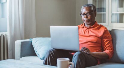 Bearded and gray African-American man in orange shirt sits with laptop.