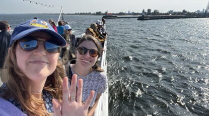 Researchers aboard a boat on the Great Lakes, wearing caps and sunglasses, wave to the camera.