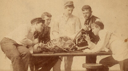 Five young men crowd around a table, on which a skeleton lies. The men are medical students of the late 19th/early 20th century. The image is sepia. A youngish man in cap gestures with a pencil while the others observe. An early anatomy ab at the University of Michigan.