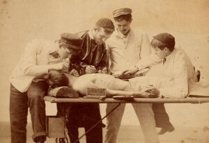 Four young men crowd around a table, on which a fifth young man lies shirtess, possibly unconscious. The men are medical students of the late 19th/early 20th century, preparing to perform a dissection. The image is sepia. A youngish man in cap lays on the table, holding th ebook open for the surgeon.