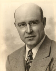 Formal headshot/sepia portrait of a balding caucasian man, mid-20th century, wearing glasses, suit, and tie.