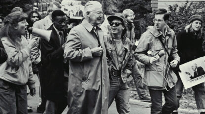 Older man in white hair and trenchcoat surrounded by protesters with bullhorns and signs. U-M campus, 1988. Robben Fleming.