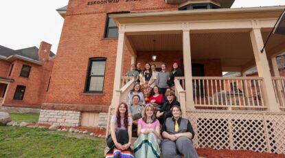 Group of people cluster together on the front porch of a red brick house. They represent the Indigenous Education Youth Collective group.