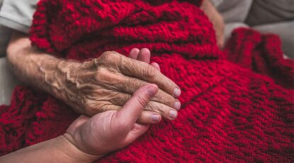 Two hands are clasped atop a red knit blanket; one is elderly, one is not. The image demonstrates the role of hospice.