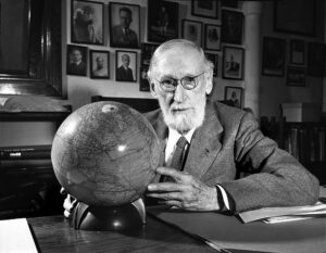 Older caucasian male, bearded and with glasses, holds a globe while sitting at a desk.