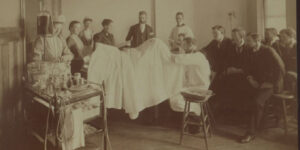 A medical school clinic in 1893 in sepia tone as University of Michigan medical students and faculty examine a body.