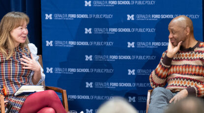 A man and woman sit on stage for an interview in front of a blue banner with white lettering that reads Gerald R. Ford School of Public Policy;