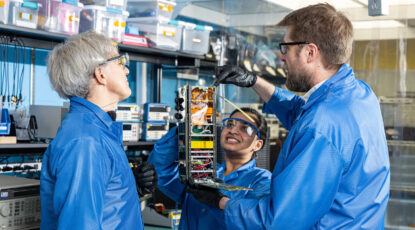 Scientists observe some kind of machine in a lab. They are wearing blue coats and protective glasses and are surrounded by machinery.