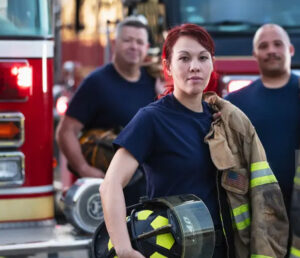 Three firefighters holding gear with truck in the background look into the camera.