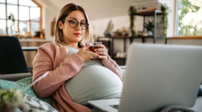 Pregnant woman with glasses holds cup of tea while leaning back on couch and looking at her laptop.