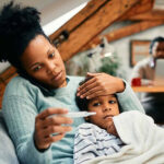 African-American mother holds young child on her lap and checks thermometer.