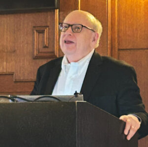 Caucasian scholar with glasses stands at podium.