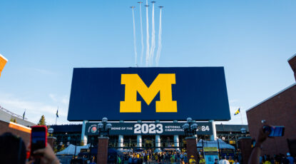 Six planes fly over Michigan Stadium, leaving white smoke trails against a bright blue sky. before the first game of the 2024 Michigan Football season. In the foreground, fans waiting to get into the gate take photos with their phones.