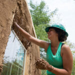 Student in green tank top and hard hat applies blobs of mud and straw to a structure in Pellston, Michigan at the U-M BioStation.