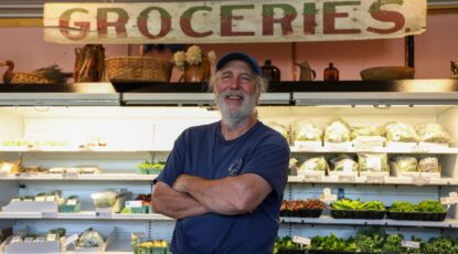 Jim Lively, co-owner of Lively NeighborFood Market in Empire, Mich., stands with arms folded in front of racks of fresh produce under a vintage painted sign that reads: Groceries.