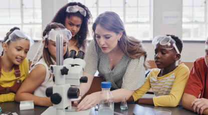 Stock image of young caucasian female teacher surrounded by students of various ethnicities, wearing safety goggles and examining a scientific instrument.