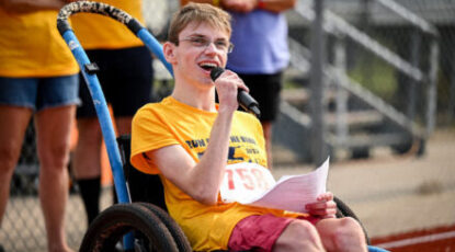 Young caucasian man in wheelchair speaks into microphone at what appears to be a competitive athletic event.