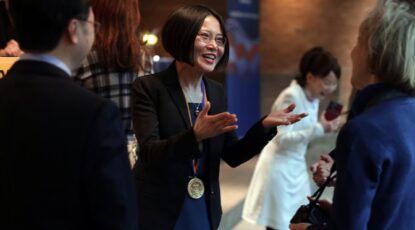Woman of Asian descent with medal around her neck, engages in animated discussion at University of Michigan event.