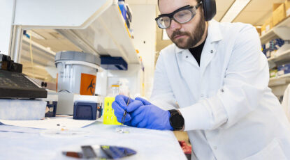 U-M scientist wearing goggles, headphones, and blue gloves, works in a laboratory on a device that could be an at-home melanoma testing patch.