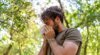 Man blows his nose, surrounded by greenery.