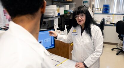 Scientist in lab coat and protective goggles points at computer screen at Michigan Medicine.