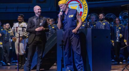 Red Hot Chili Peppers drummer Chad Smith on stage with the Michigan Marching Band at Hill Auditorium in Ann Arbor. Band is in uniform. Smith wears a blue jumpsuit with maize baseball cap turned backwards.