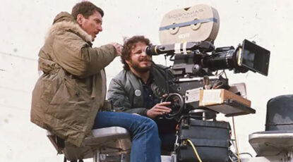 Film director Lawrence Kasdan prepares to film a shot by looking through the lens of a Panavision camera while another filmmaker dressed in a parka reviews.