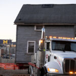 Massive semi pulls a house from its original foundation in Ann Arbor to a new location. University of Michigan stadium is in the background.