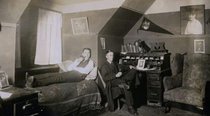 Black and white photo of two male University of Michigan students in their dorm room circa 1920. Lounging and smoking a pipe.