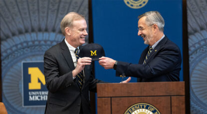 Two men stand at podium, holding a ball cap with University of Michigan Block M.