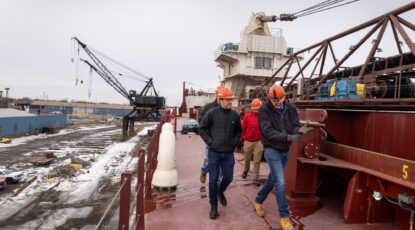 Men in hardhats walk along the deck of a ship in what appears to be a major shipping yard.